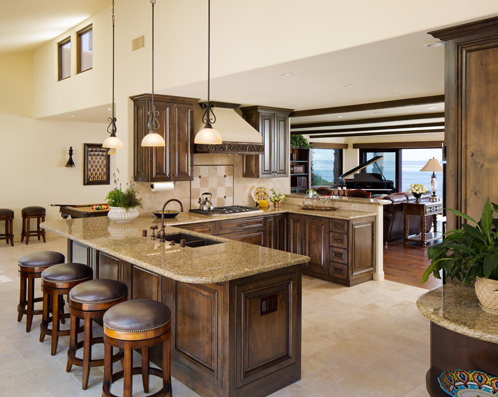 Example of a mid-sized tuscan u-shaped ceramic tile open concept kitchen design in San Diego with raised-panel cabinets, dark wood cabinets, beige backsplash, a peninsula, an undermount sink, granite countertops, porcelain backsplash and paneled appliances