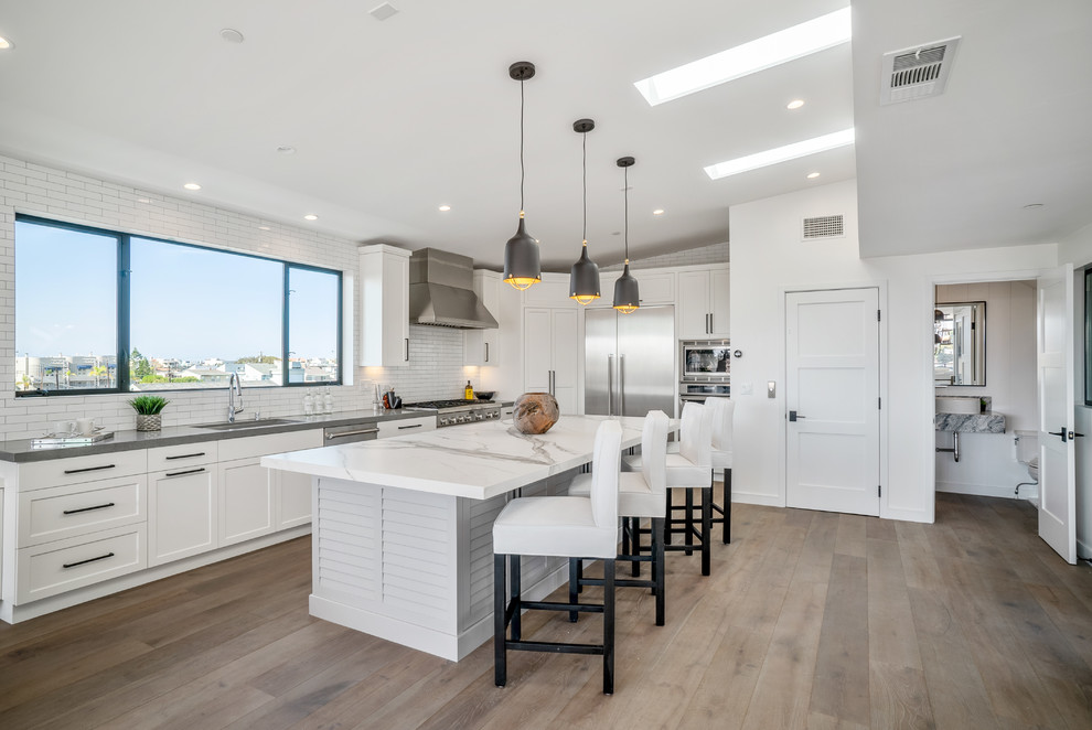 Inspiration for a classic l-shaped kitchen in Los Angeles with a submerged sink, shaker cabinets, white cabinets, white splashback, metro tiled splashback, stainless steel appliances, medium hardwood flooring, an island, brown floors and white worktops.