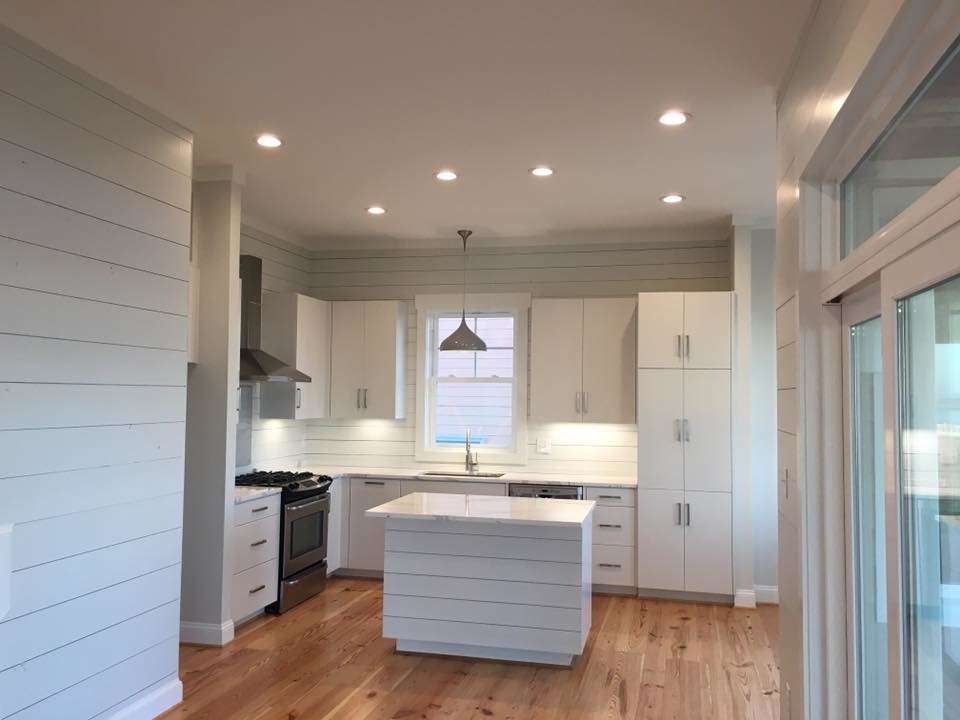 This is an example of a beach style l-shaped kitchen in Other with flat-panel cabinets, white cabinets, quartz worktops, white splashback, wood splashback, stainless steel appliances, medium hardwood flooring and an island.