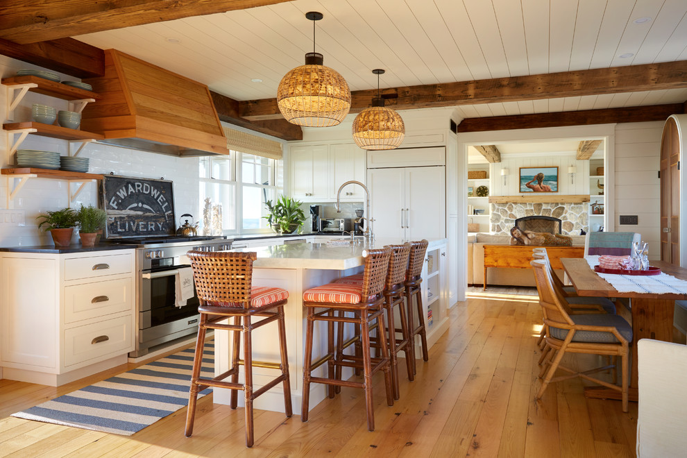 Design ideas for a coastal l-shaped kitchen/diner in Portland Maine with shaker cabinets, white cabinets, white splashback, integrated appliances, light hardwood flooring, an island and black worktops.