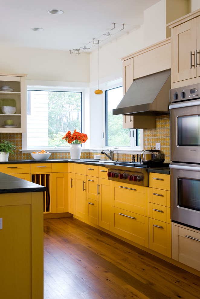 Ejemplo de cocina tradicional con armarios estilo shaker, puertas de armario amarillas, salpicadero amarillo, electrodomésticos de acero inoxidable, suelo de madera en tonos medios y una isla