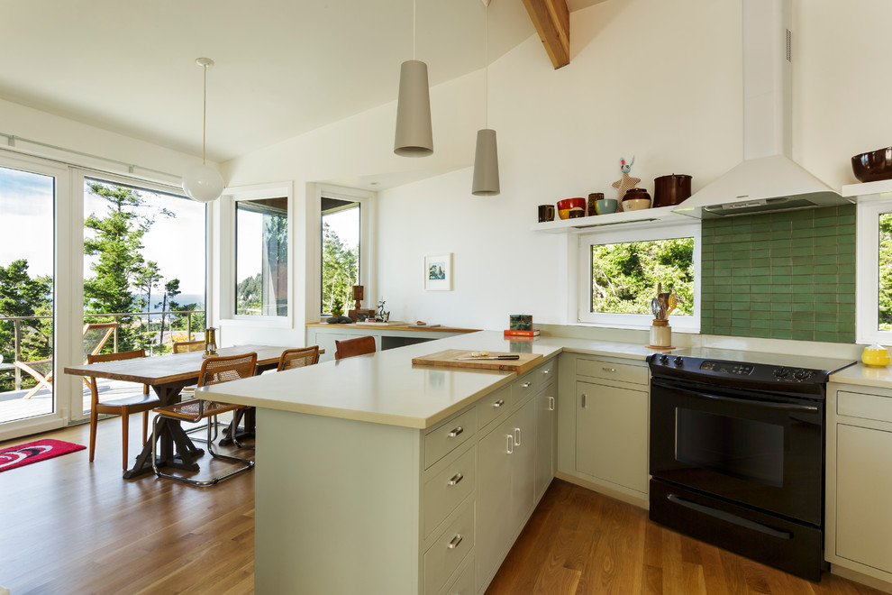 Offene Maritime Küche mit flächenbündigen Schrankfronten, beigen Schränken, Küchenrückwand in Grün, schwarzen Elektrogeräten und braunem Holzboden in Portland