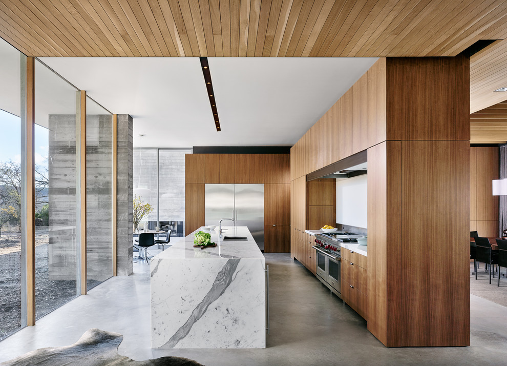 Cette photo montre une grande cuisine ouverte moderne en bois brun avec un placard à porte plane, îlot, un évier encastré, un électroménager en acier inoxydable, sol en béton ciré, un sol gris et un plan de travail blanc.
