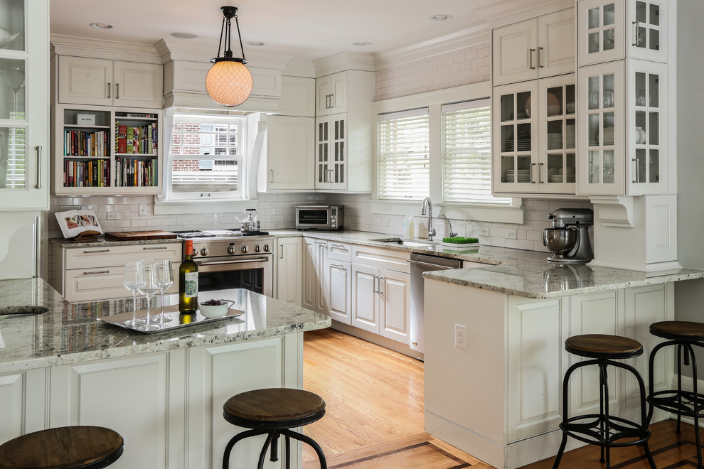 Exempel på ett klassiskt kök, med en undermonterad diskho, luckor med glaspanel, vita skåp, vitt stänkskydd, stänkskydd i tunnelbanekakel, rostfria vitvaror, mellanmörkt trägolv och en halv köksö