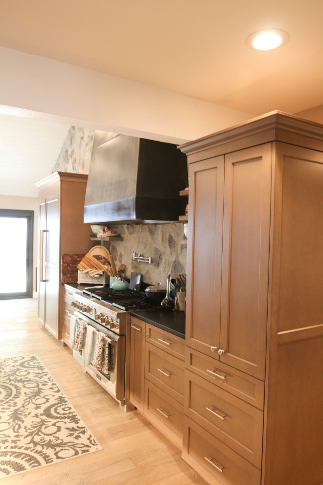 This is an example of a medium sized country u-shaped open plan kitchen in Los Angeles with a belfast sink, brown cabinets, black splashback, stone tiled splashback, stainless steel appliances, medium hardwood flooring, an island, multicoloured worktops and a vaulted ceiling.