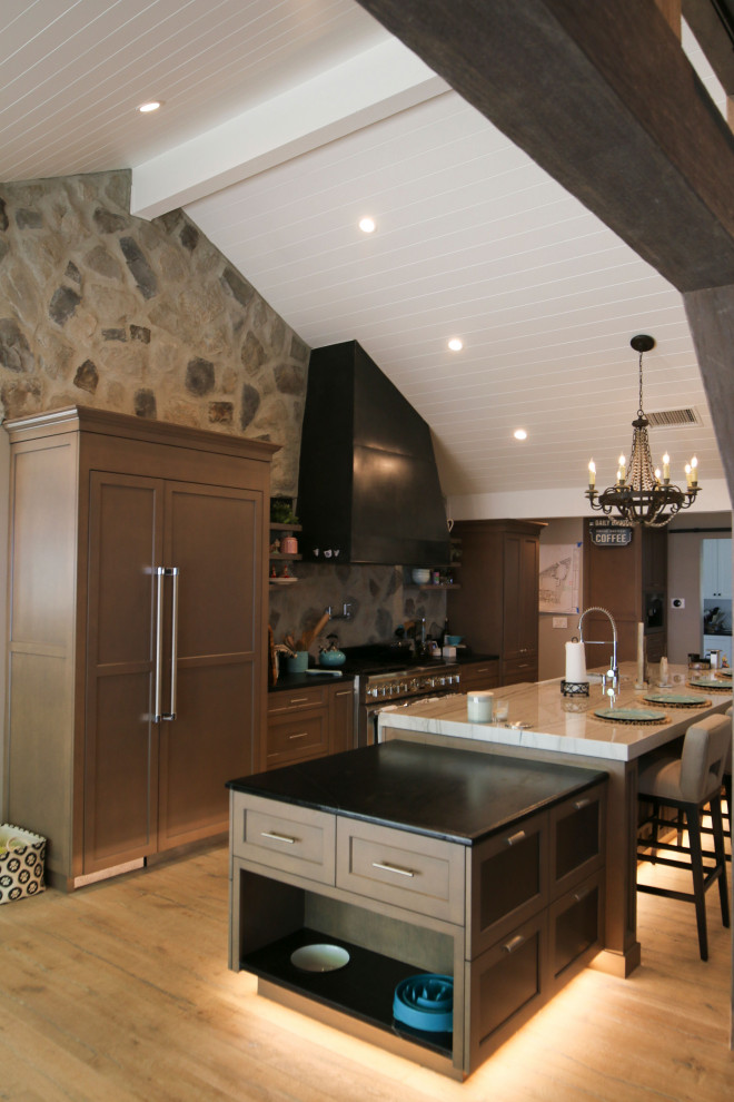 Medium sized farmhouse u-shaped open plan kitchen in Los Angeles with a belfast sink, brown cabinets, black splashback, stone tiled splashback, stainless steel appliances, medium hardwood flooring, an island, multicoloured worktops and a vaulted ceiling.