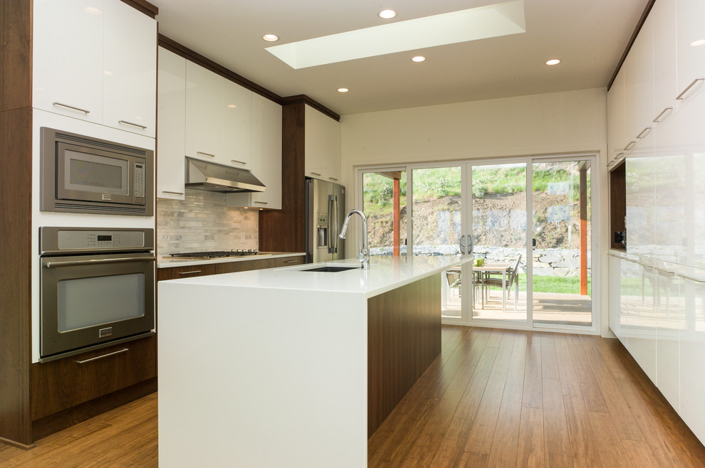 Bild på ett avskilt, mellanstort funkis linjärt kök, med en undermonterad diskho, släta luckor, vita skåp, bänkskiva i kvarts, grått stänkskydd, stänkskydd i stenkakel, rostfria vitvaror, ljust trägolv, en köksö och beiget golv