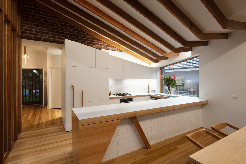 Small contemporary u-shaped kitchen/diner in Melbourne with white cabinets, laminate countertops, white splashback, ceramic splashback, medium hardwood flooring, brown floors, white worktops, flat-panel cabinets, integrated appliances and a breakfast bar.