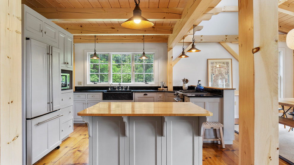 This is an example of a large rural l-shaped open plan kitchen in New York with a belfast sink, recessed-panel cabinets, white cabinets, soapstone worktops, stainless steel appliances, medium hardwood flooring, an island, black worktops and exposed beams.