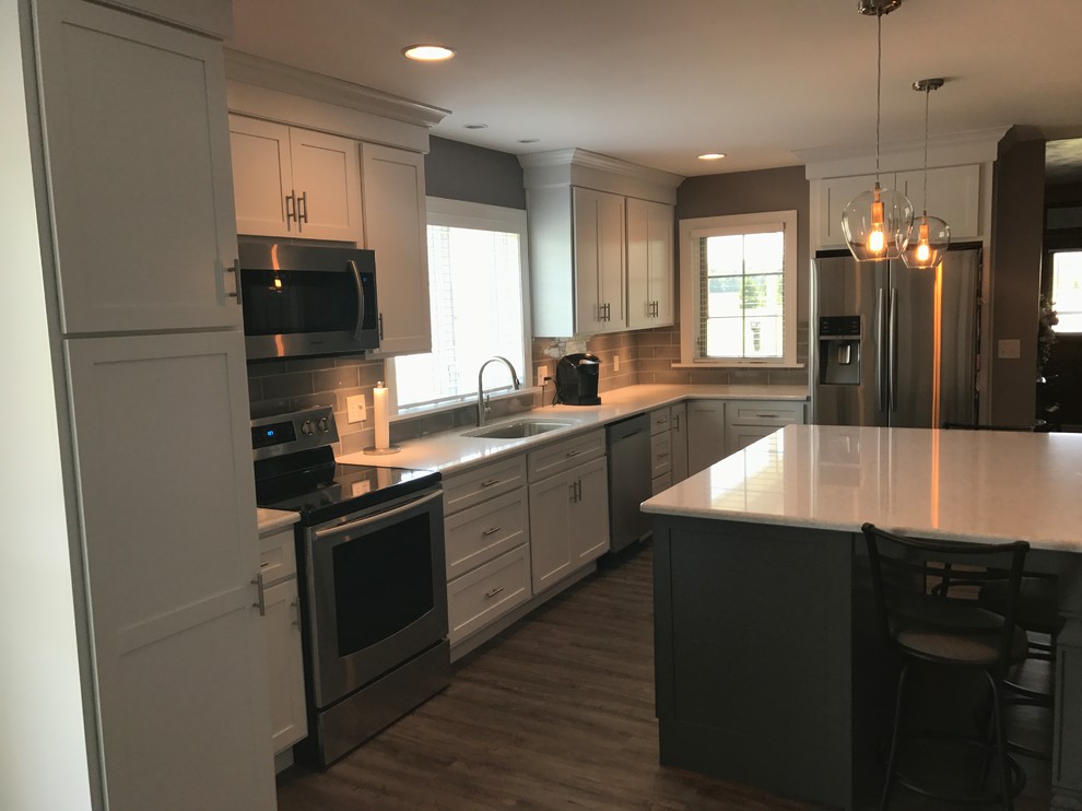 Photo of an expansive country l-shaped kitchen pantry in Other with a submerged sink, shaker cabinets, white cabinets, engineered stone countertops, grey splashback, stainless steel appliances, an island and brown floors.
