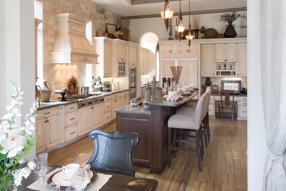 Example of a large classic single-wall medium tone wood floor and brown floor enclosed kitchen design in Miami with paneled appliances, a farmhouse sink, raised-panel cabinets, distressed cabinets, granite countertops, brown backsplash, travertine backsplash and an island