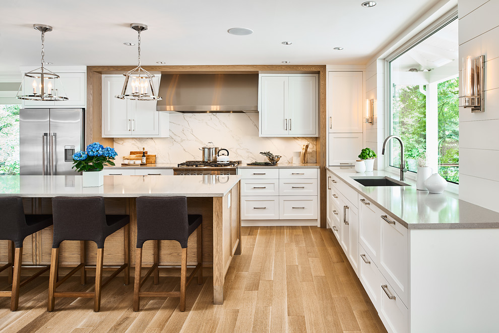 Klassische Küche in L-Form mit Unterbauwaschbecken, Schrankfronten im Shaker-Stil, weißen Schränken, Rückwand-Fenster, Küchengeräten aus Edelstahl, hellem Holzboden, Kücheninsel und grauer Arbeitsplatte in Vancouver