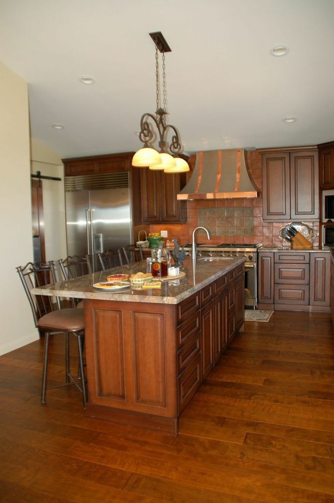 Cette photo montre une cuisine américaine chic en L et bois brun de taille moyenne avec un placard avec porte à panneau surélevé, un plan de travail en granite, une crédence orange, une crédence en carreau de porcelaine, un électroménager en acier inoxydable, un sol en bois brun et îlot.