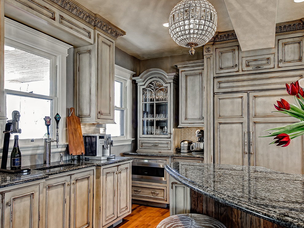 Example of a large classic light wood floor kitchen design in Denver with an undermount sink, beaded inset cabinets, distressed cabinets, granite countertops, green backsplash, glass tile backsplash, paneled appliances and an island