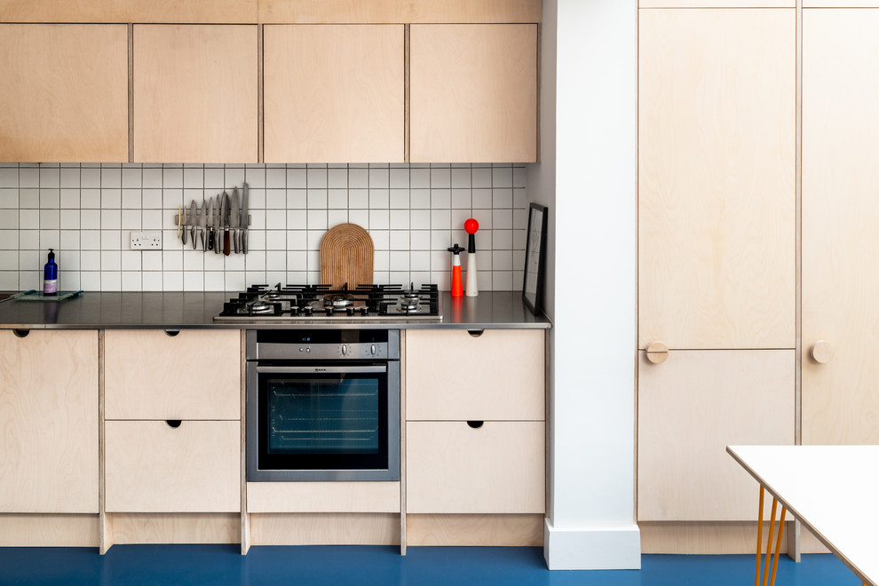 ロンドンにある北欧スタイルのおしゃれなキッチン (淡色木目調キャビネット、ステンレスカウンター、青い床、フラットパネル扉のキャビネット、白いキッチンパネル、シルバーの調理設備) の写真