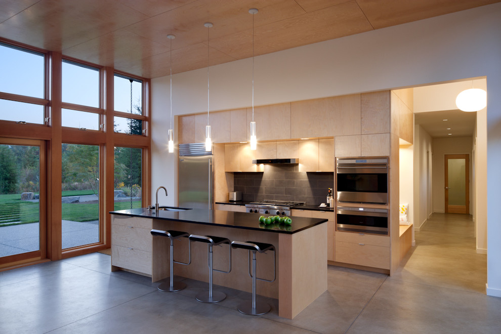 Cette image montre une cuisine parallèle minimaliste en bois clair avec un placard à porte plane, une crédence noire, un électroménager en acier inoxydable et une crédence en ardoise.