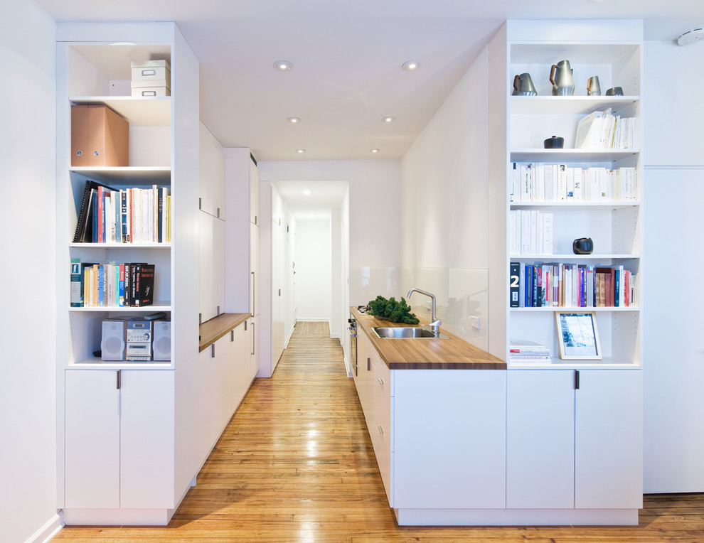 Contemporary single-wall kitchen pantry in New York with a built-in sink, flat-panel cabinets, white cabinets, wood worktops, white splashback, stainless steel appliances and medium hardwood flooring.