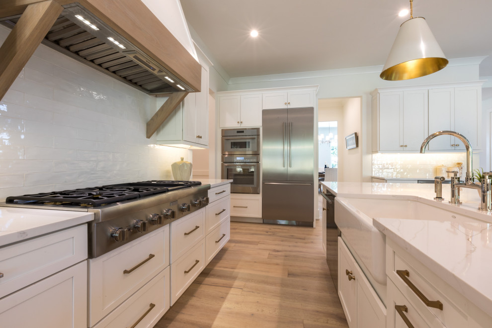 Inspiration for a mid-sized coastal u-shaped medium tone wood floor and brown floor eat-in kitchen remodel in Orlando with a farmhouse sink, shaker cabinets, white cabinets, quartz countertops, white backsplash, ceramic backsplash, stainless steel appliances, an island and white countertops