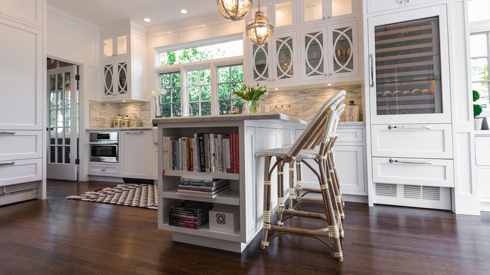 Cette photo montre une cuisine américaine chic de taille moyenne avec un placard à porte shaker, des portes de placard blanches, un plan de travail en granite, une crédence beige, une crédence en carrelage de pierre, parquet foncé, une péninsule et un sol marron.