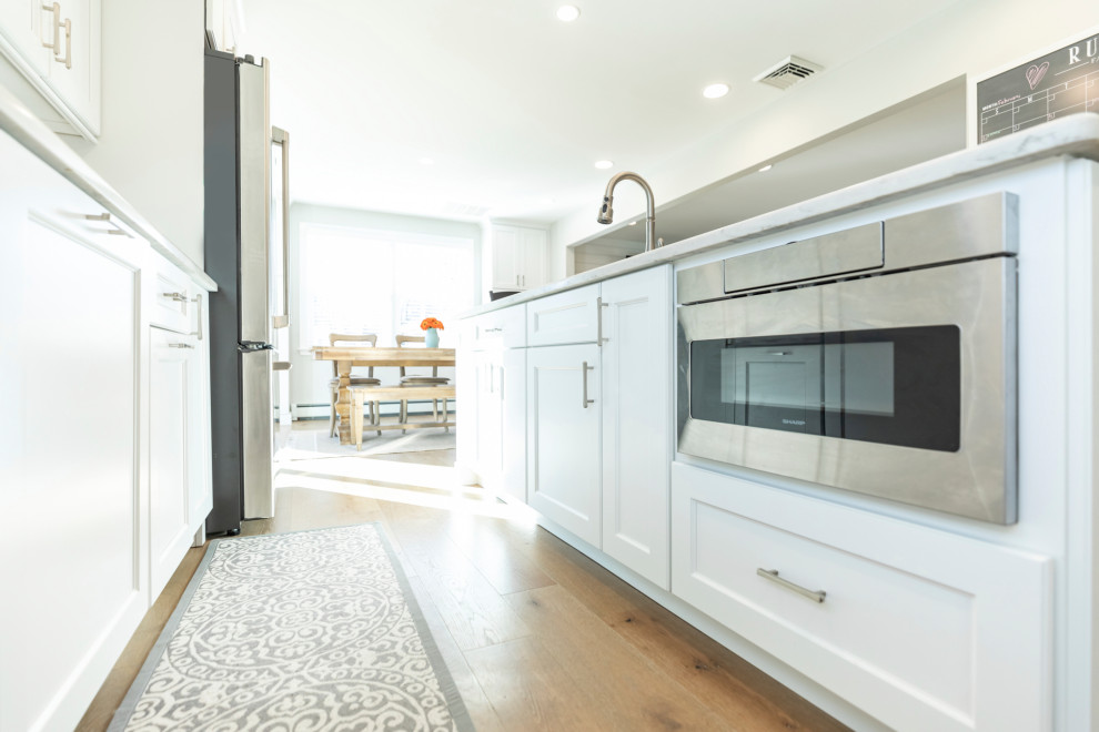 Photo of a medium sized traditional l-shaped open plan kitchen in Philadelphia with a submerged sink, recessed-panel cabinets, white cabinets, quartz worktops, white splashback, porcelain splashback, stainless steel appliances, medium hardwood flooring, an island, brown floors and beige worktops.
