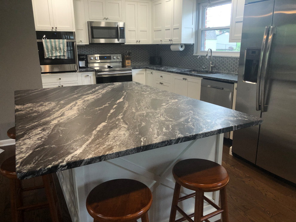 Photo of a large traditional l-shaped kitchen/diner in Indianapolis with a single-bowl sink, shaker cabinets, white cabinets, granite worktops, grey splashback, porcelain splashback, stainless steel appliances, medium hardwood flooring, an island, brown floors and black worktops.