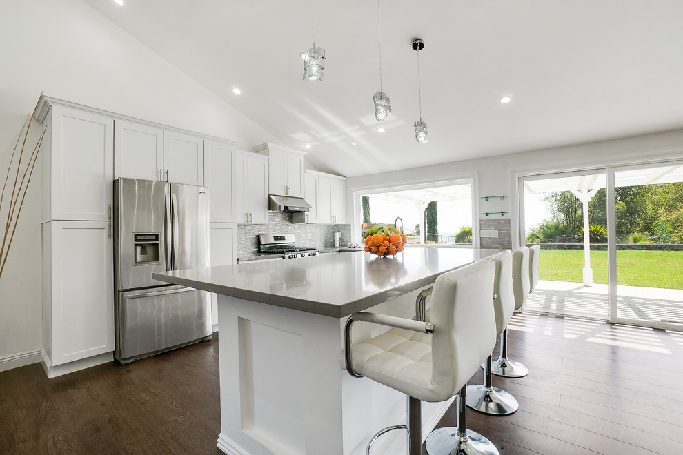 Inspiration for a large modern l-shaped kitchen/diner in Seattle with a belfast sink, shaker cabinets, white cabinets, engineered stone countertops, grey splashback, glass sheet splashback, stainless steel appliances, laminate floors, an island, brown floors and grey worktops.