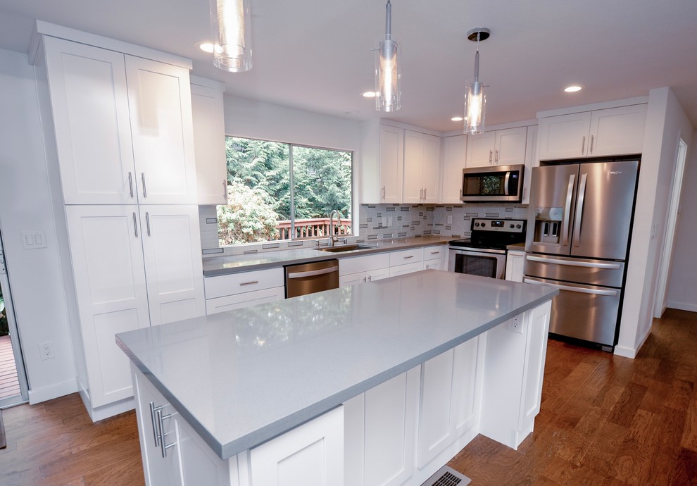 Large minimalist l-shaped brown floor and laminate floor kitchen photo in Seattle with shaker cabinets, white cabinets, quartzite countertops, stainless steel appliances, an island, gray countertops, an integrated sink, gray backsplash and ceramic backsplash