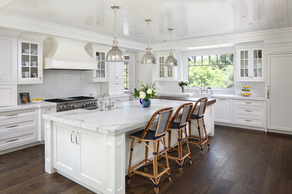 Inspiration for a coastal l-shaped kitchen in San Francisco with a belfast sink, shaker cabinets, white cabinets, white splashback, metro tiled splashback, integrated appliances, dark hardwood flooring, an island, brown floors and white worktops.
