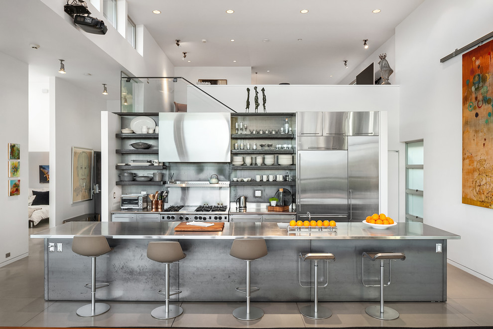 Bild på ett mycket stort funkis grå grått kök, med en integrerad diskho, släta luckor, skåp i rostfritt stål, bänkskiva i rostfritt stål, stänkskydd med metallisk yta, stänkskydd i metallkakel, rostfria vitvaror, betonggolv, en köksö och grått golv