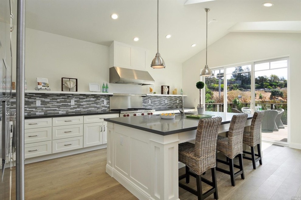 This is an example of a large coastal single-wall kitchen/diner in San Francisco with a submerged sink, shaker cabinets, white cabinets, composite countertops, multi-coloured splashback, matchstick tiled splashback, stainless steel appliances, light hardwood flooring and an island.