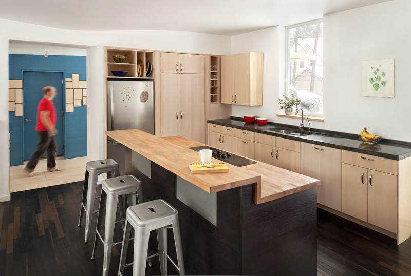 На фото: параллельная кухня-гостиная в современном стиле с двойной мойкой, плоскими фасадами, светлыми деревянными фасадами, техникой из нержавеющей стали, деревянным полом и островом