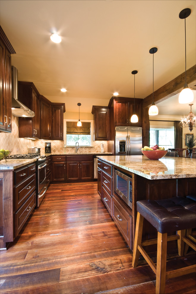 Example of a mid-sized mountain style l-shaped medium tone wood floor open concept kitchen design in New Orleans with an undermount sink, raised-panel cabinets, dark wood cabinets, granite countertops, beige backsplash, stone tile backsplash, stainless steel appliances and an island