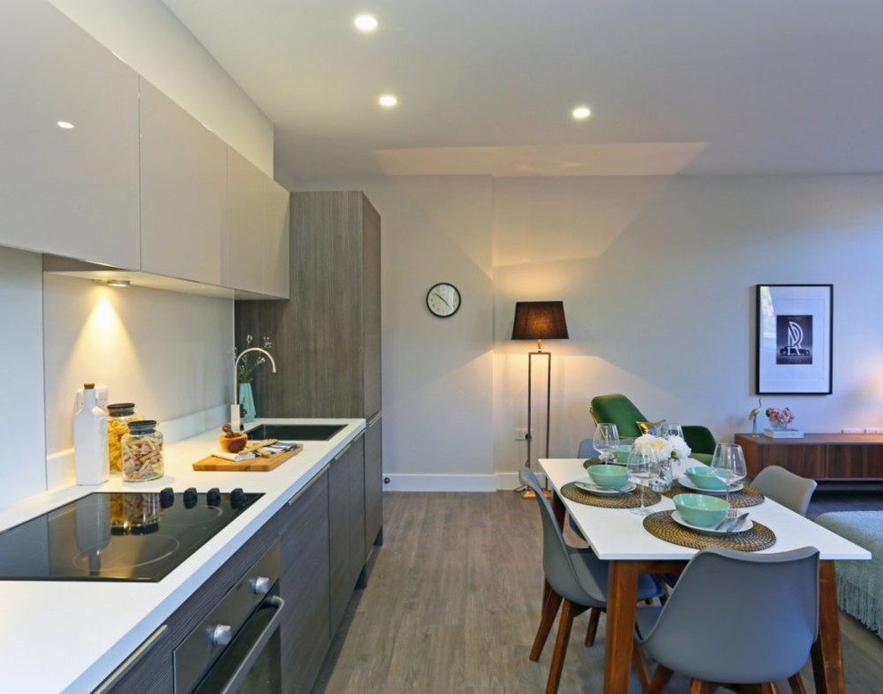 This is an example of a small modern single-wall kitchen/diner in Cheshire with a single-bowl sink, flat-panel cabinets, dark wood cabinets, composite countertops, beige splashback, glass sheet splashback, black appliances, medium hardwood flooring, no island, brown floors and white worktops.