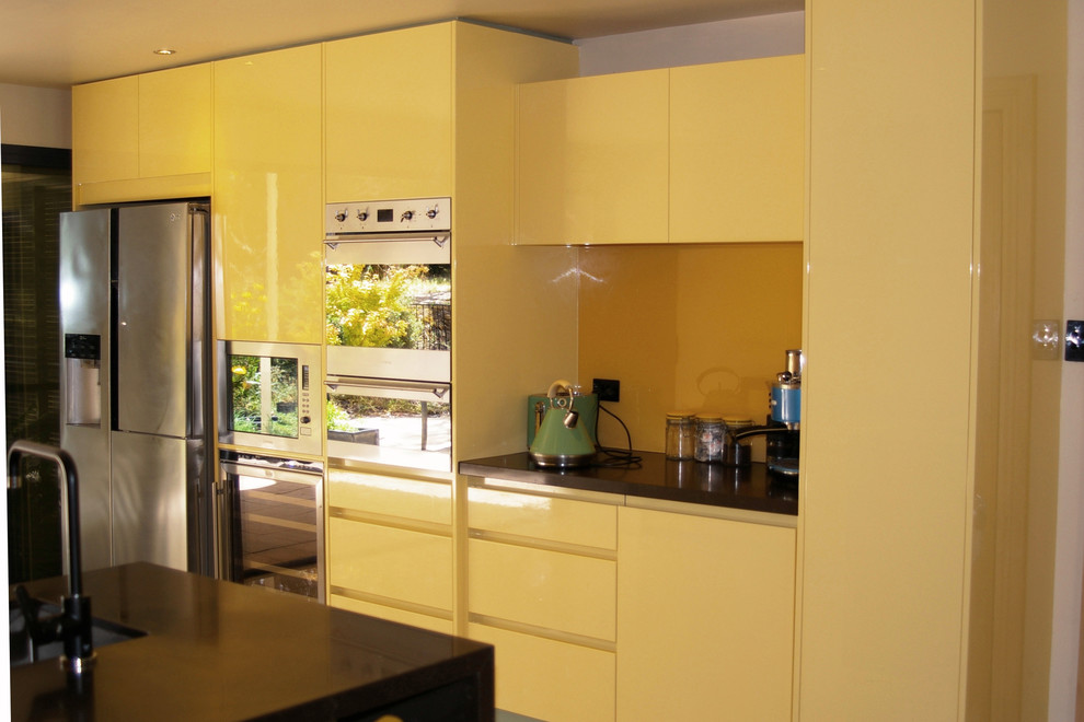 Идея дизайна: угловая кухня-гостиная среднего размера в современном стиле с врезной мойкой, плоскими фасадами, желтыми фасадами, столешницей из кварцевого агломерата, желтым фартуком, фартуком из стекла, техникой из нержавеющей стали, полом из керамогранита, полуостровом и бежевым полом