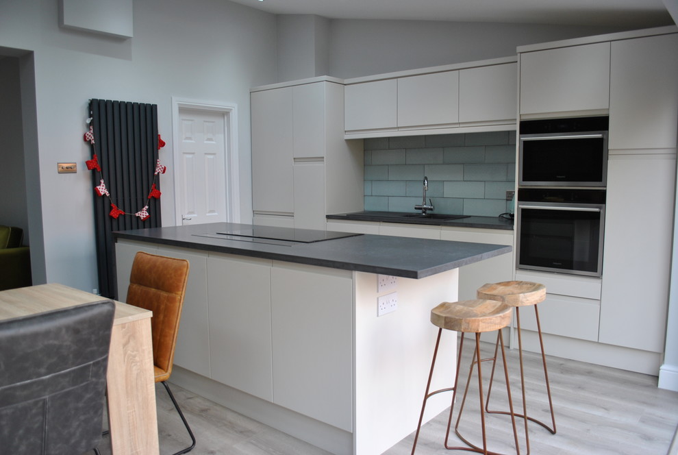 Design ideas for a medium sized contemporary kitchen/diner in Hertfordshire with a submerged sink, flat-panel cabinets, metro tiled splashback, black appliances, laminate floors, an island and grey floors.