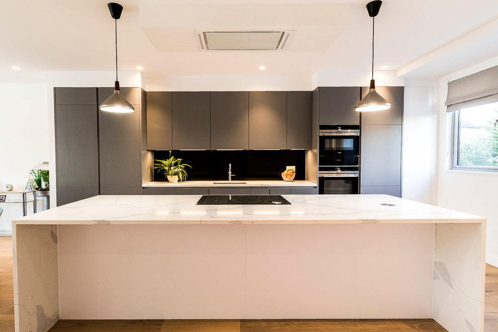 Foto de cocina lineal actual de tamaño medio abierta con puertas de armario grises, encimera de acrílico, salpicadero negro, salpicadero de vidrio templado y una isla