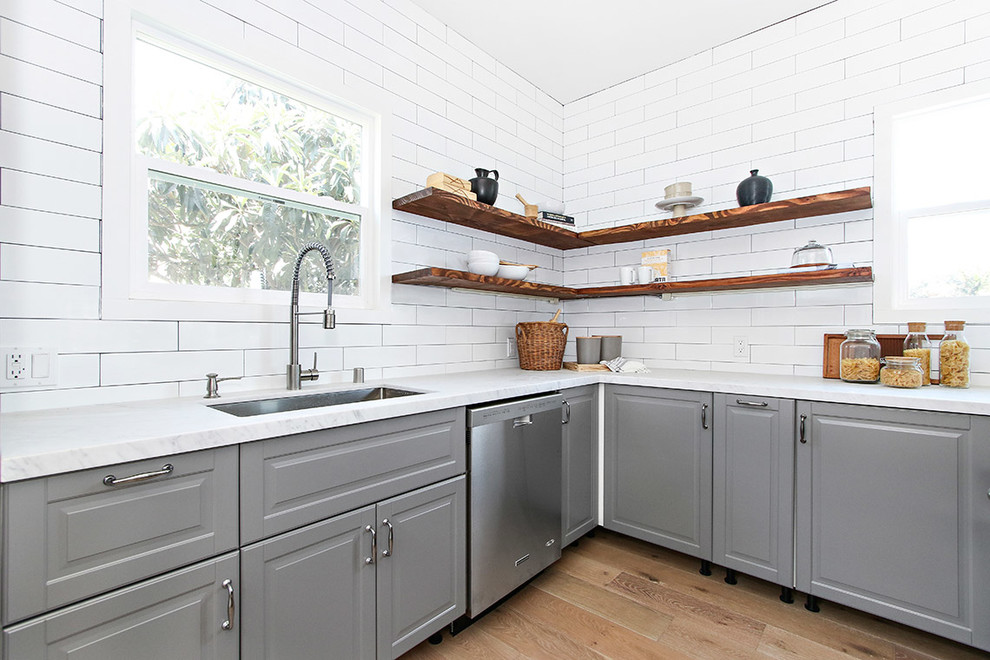 Bild på ett vintage l-kök, med en undermonterad diskho, luckor med upphöjd panel, grå skåp, marmorbänkskiva, vitt stänkskydd, stänkskydd i tunnelbanekakel, rostfria vitvaror och ljust trägolv