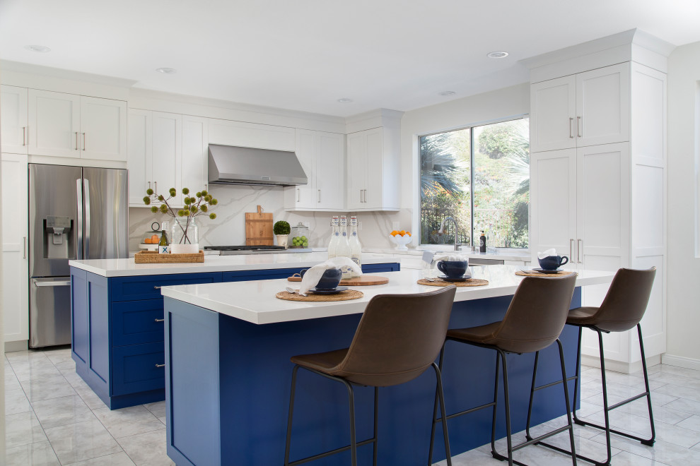 Cette photo montre une cuisine chic en L de taille moyenne avec un placard à porte shaker, des portes de placard bleues, un électroménager en acier inoxydable, 2 îlots, un sol gris et un plan de travail blanc.
