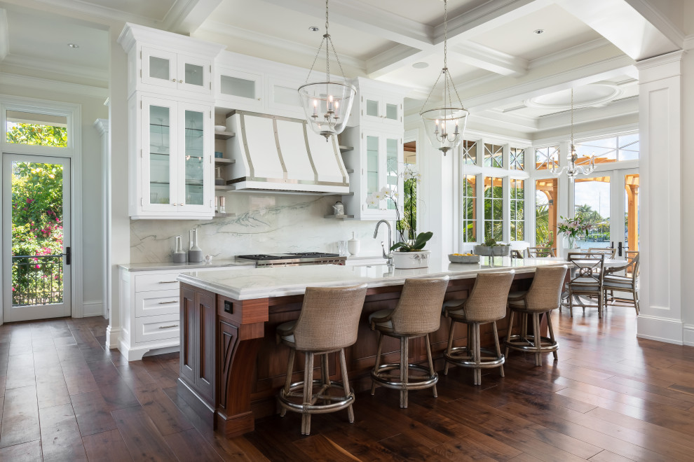 Photo of a large beach style single-wall kitchen/diner in Tampa with a belfast sink, glass-front cabinets, white cabinets, marble worktops, marble splashback, stainless steel appliances, dark hardwood flooring, an island, brown floors and white worktops.