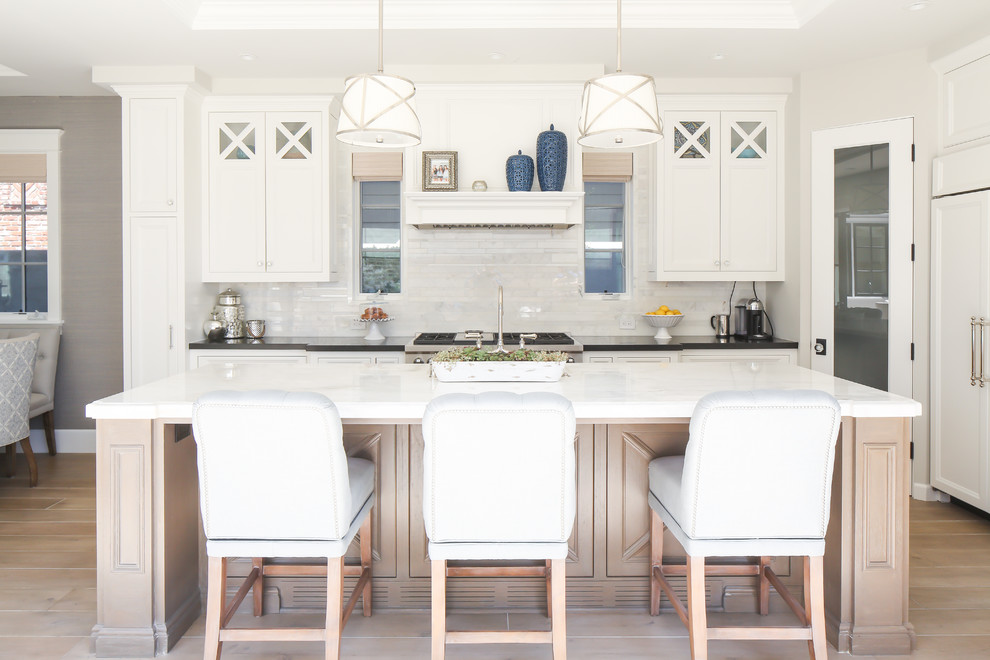 Diseño de cocinas en L costera con puertas de armario blancas, salpicadero blanco, electrodomésticos con paneles, suelo de madera clara y una isla