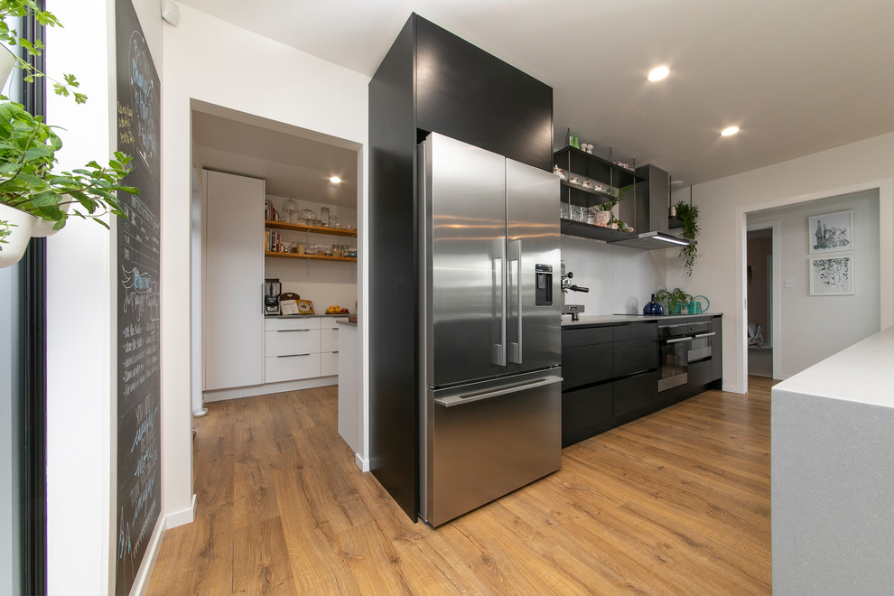 Photo of a large contemporary galley kitchen/diner in Auckland with a single-bowl sink, flat-panel cabinets, black cabinets, engineered stone countertops, white splashback, stainless steel appliances, laminate floors, an island, brown floors and grey worktops.