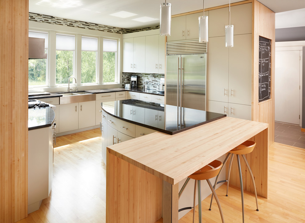 Trendy u-shaped medium tone wood floor kitchen photo in Minneapolis with a farmhouse sink, flat-panel cabinets, white cabinets, multicolored backsplash, stainless steel appliances and an island