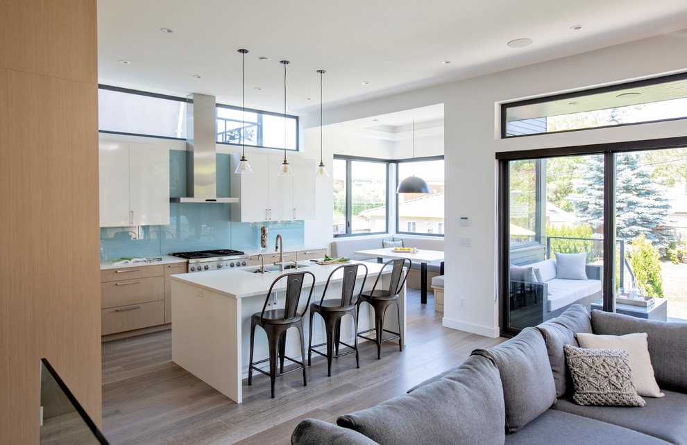 Photo of a large contemporary l-shaped open plan kitchen in Vancouver with a double-bowl sink, flat-panel cabinets, light wood cabinets, engineered stone countertops, blue splashback, glass sheet splashback, stainless steel appliances, medium hardwood flooring, an island, grey floors and white worktops.