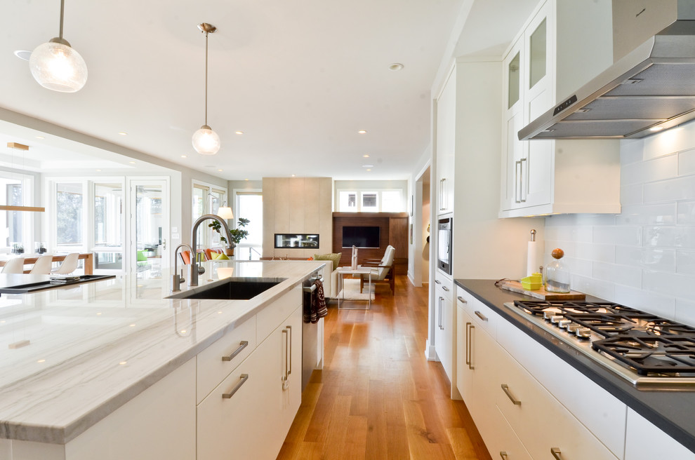 Modern inredning av ett kök med öppen planlösning, med släta luckor, stänkskydd i glaskakel, rostfria vitvaror, en undermonterad diskho, vitt stänkskydd och vita skåp