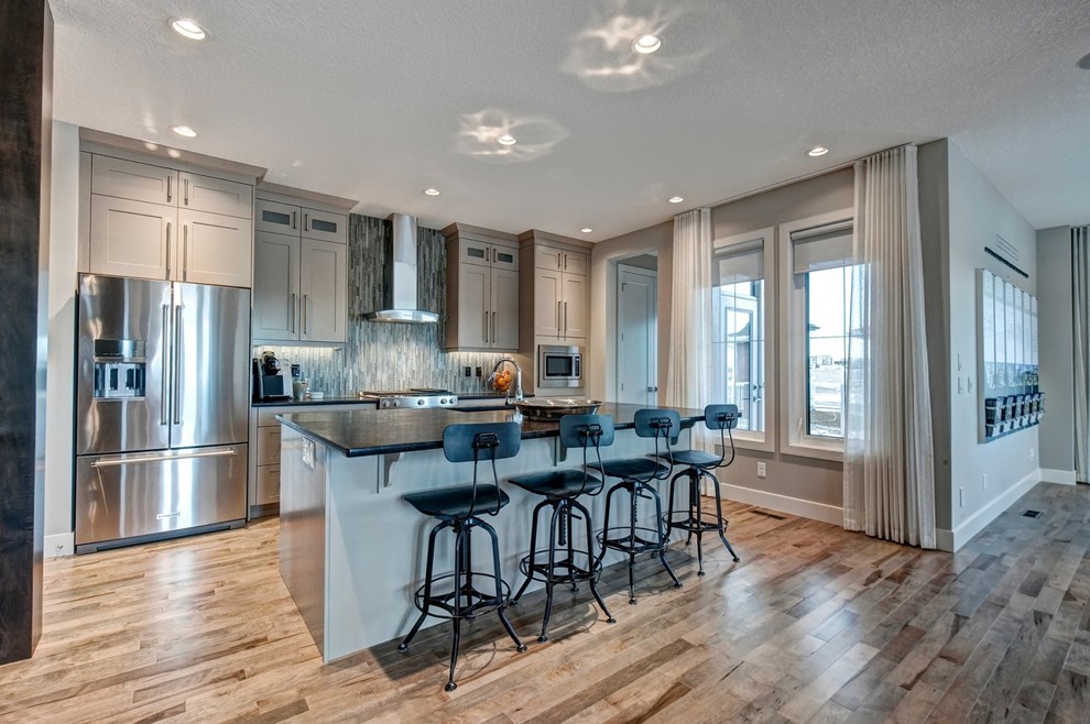 This is an example of a medium sized contemporary l-shaped open plan kitchen in Calgary with a submerged sink, recessed-panel cabinets, grey cabinets, quartz worktops, grey splashback, glass tiled splashback, stainless steel appliances, medium hardwood flooring, an island and brown floors.