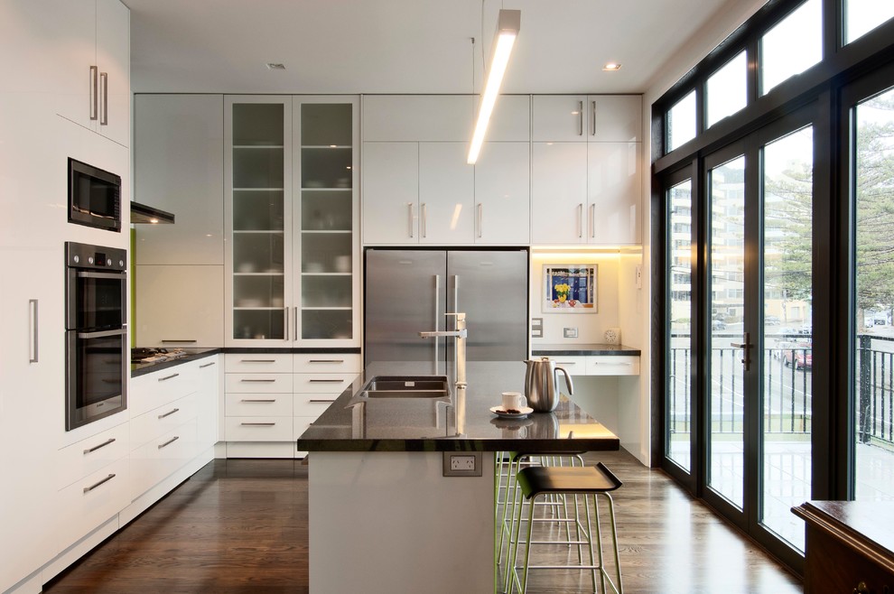 Photo of a contemporary l-shaped open plan kitchen in Auckland with a submerged sink, flat-panel cabinets, white cabinets, green splashback, glass sheet splashback, stainless steel appliances, dark hardwood flooring and an island.