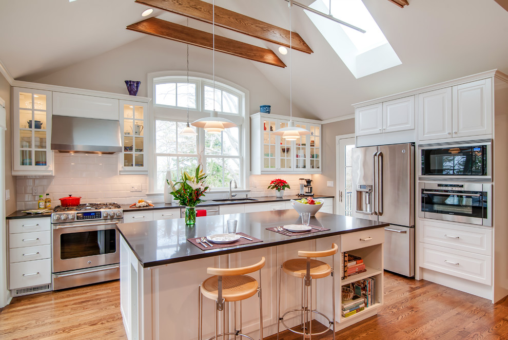 Photo of a traditional kitchen in New York with a submerged sink, glass-front cabinets, white cabinets, white splashback, metro tiled splashback, stainless steel appliances, medium hardwood flooring and an island.