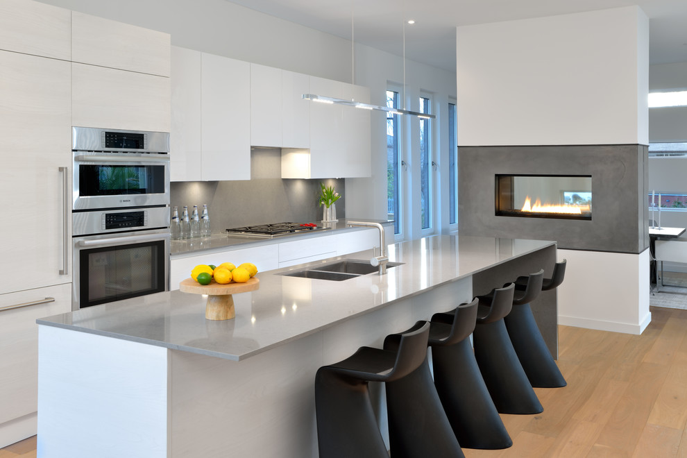 Contemporary galley kitchen in Toronto with a submerged sink, flat-panel cabinets, white cabinets, grey splashback, stainless steel appliances, an island and light hardwood flooring.