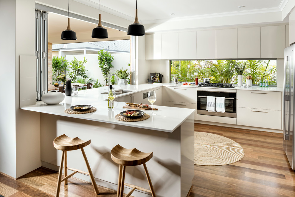Inredning av ett modernt kök, med en dubbel diskho, släta luckor, beige skåp, fönster som stänkskydd, rostfria vitvaror, mellanmörkt trägolv, en halv köksö och brunt golv