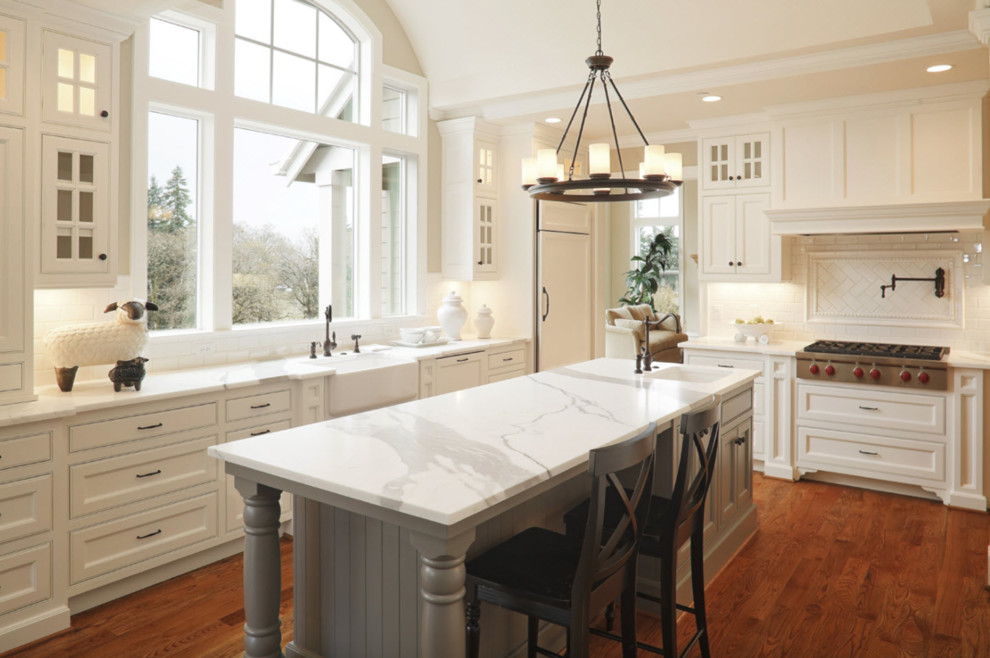 Lantlig inredning av ett avskilt, stort vit vitt l-kök, med en rustik diskho, luckor med infälld panel, vita skåp, marmorbänkskiva, vitt stänkskydd, stänkskydd i tunnelbanekakel, integrerade vitvaror, mörkt trägolv, en köksö och brunt golv